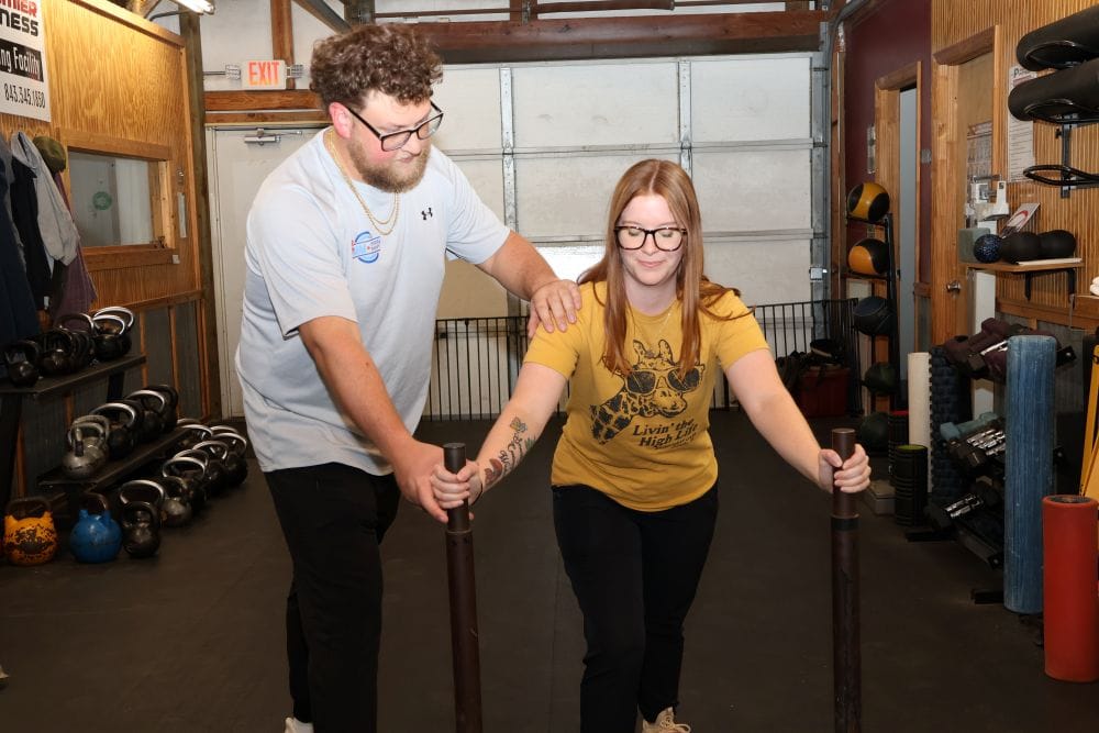 AIMPT - A man assists a woman as she pushes weighted sled bars in a gym setting with kettlebells and equipment visible in the background. - Charleston, South Carolina