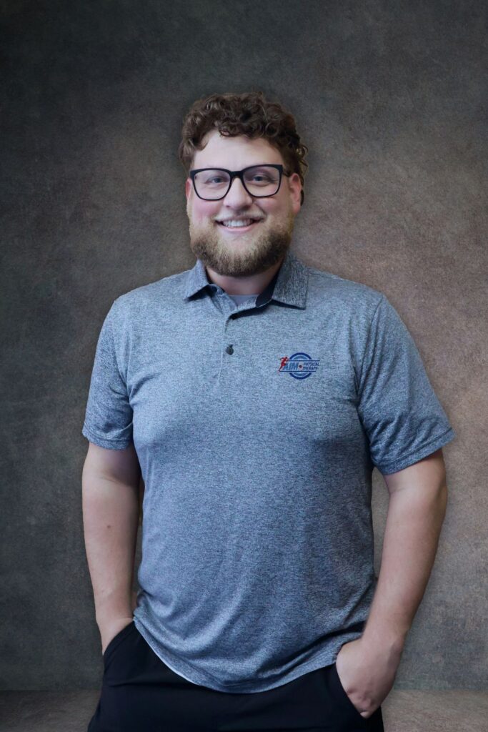 AIMPT - A man with curly hair, glasses, and a beard stands smiling with hands in pockets, wearing a gray polo shirt against a neutral studio background. - Charleston, South Carolina