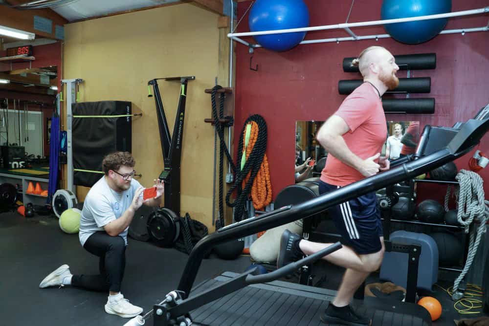 AIMPT - A man exercises on a treadmill while another man kneels nearby, holding a stopwatch and observing him in a gym setting. - Charleston, South Carolina