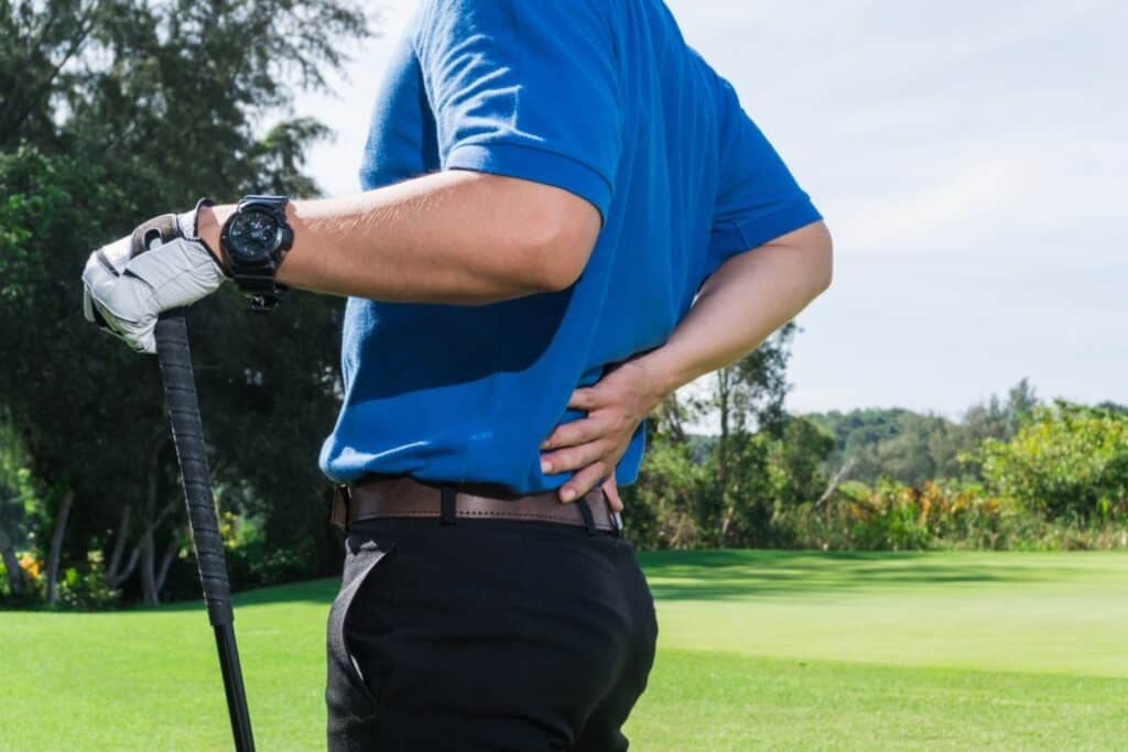 AIMPT - A golfer in a blue shirt and black pants holds his lower back with one hand and supports himself with a golf club, standing on a golf course. - Charleston, South Carolina
